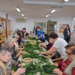 Skupina starejših oseb in skrbnikov, ki skupaj izdelujejo adventne venčke z uporabo zelenja in slame, med delom za mizo.