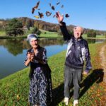 Starejši moški in ženska stojita ob reki, moški veselo meče jesensko listje v zrak, medtem ko ženska gleda z nasmeškom. Jesenska pokrajina z jezerom in zelenjem v ozadju.