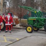 Šest ljudi v božičkovih kostumih stoji ob dveh traktorjih, enem rdečem in enem zelenem. Scena je postavljena na parkirišču, v ozadju so stavbe in drevesa. Božički mahajo in so nasmejani.