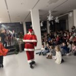 Fotografija prikazuje Božička, ki hodi proti skupini otrok, oblečen v tradicionalno rdečo opravo z belim krznom. Otroci sedijo na tleh na blazinah in z navdušenjem spremljajo njegov prihod. V ospredju je več deklic in dečkov, med njimi deklica v beli obleki, ki pozorno gleda. Prostor je okrašen s snežinkami, ki visijo s stropa, in papirnatimi božičnimi smrekami na oknih. Praznična dvorana je polna otrok, staršev in prijetnega vzdušja.