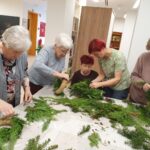 Pet žensk stoji za mizo in natančno oblikuje adventne venčke iz zelenih vej in naravnih materialov. Na mizi je videti razporejene veje in orodje.