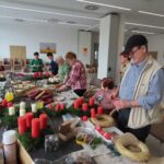 Večje število ljudi ustvarja adventne venčke okoli dolge mize. Na mizi so položene sveče, storži, trakovi in drugi okraski za venčke.