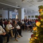 Soba polna ljudi, ki sedijo na stolih in poslušajo dogodek. V ospredju je božično drevo okrašeno z lučkami in okraski. V ozadju so obešene bele papirnate snežinke.