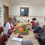 Skupina starejših žensk in skrbnica sedijo za dolgo leseno mizo. Na mizi so razpršeni materiali za ustvarjanje, vključno z volno v različnih barvah, škarjami in kartonskimi tulci. Skupaj ustvarjajo in se družijo v prijetnem in svetlem prostoru.