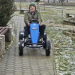 Starejši moški v zeleni bundi in temni pleteni kapi vozi moder gokart po tlakovani poti. Pogleda naravnost naprej in drži volan. Ob poti so betonske klopi, nizka kovinska ograja in majhni grmički, prekriti s tanko plastjo snega.