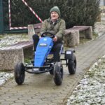 Moški v zeleni bundi in pleteni kapi se pelje z modrim gokartom po potki, obdani z zasneženo travo. Ob njem je lesena klop z betonskimi nogami, v ozadju pa visoka zelena živa meja in rdeče-belo označena vrata. Pogleda naravnost naprej in drži volan.